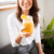 Smiling woman holding a mason jar of fresh orange juice with orange slice garnish indoors.
